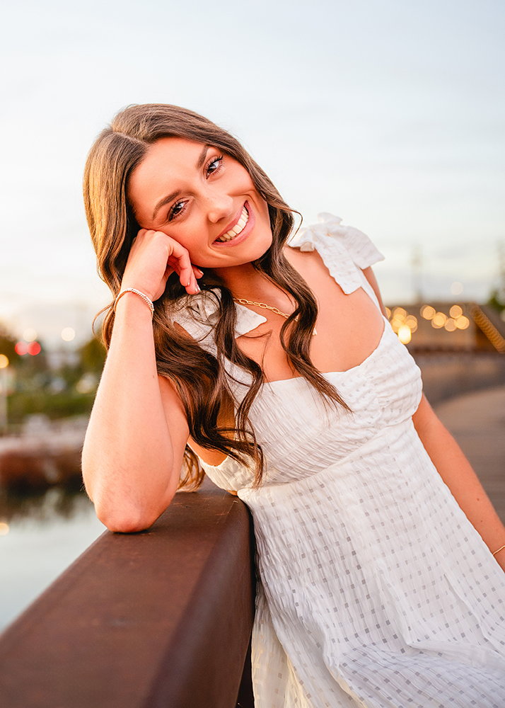 A high school senior in a white dress leans on a wooden railing, smiling warmly. The background features a softly blurred evening sky and glowing bokeh lights, creating a serene and joyful atmosphere.
