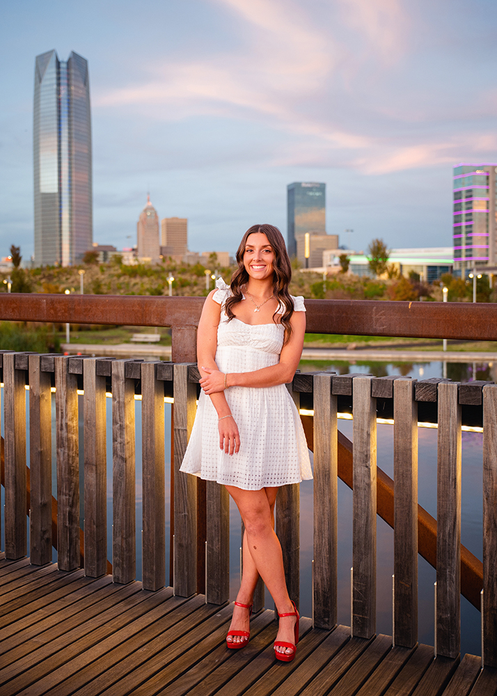Macey poses for her senior photos in a white dress and red heels on a wooden bridge at sunset, Oklahoma City skyscrapers in the background; warm, cheerful atmosphere.