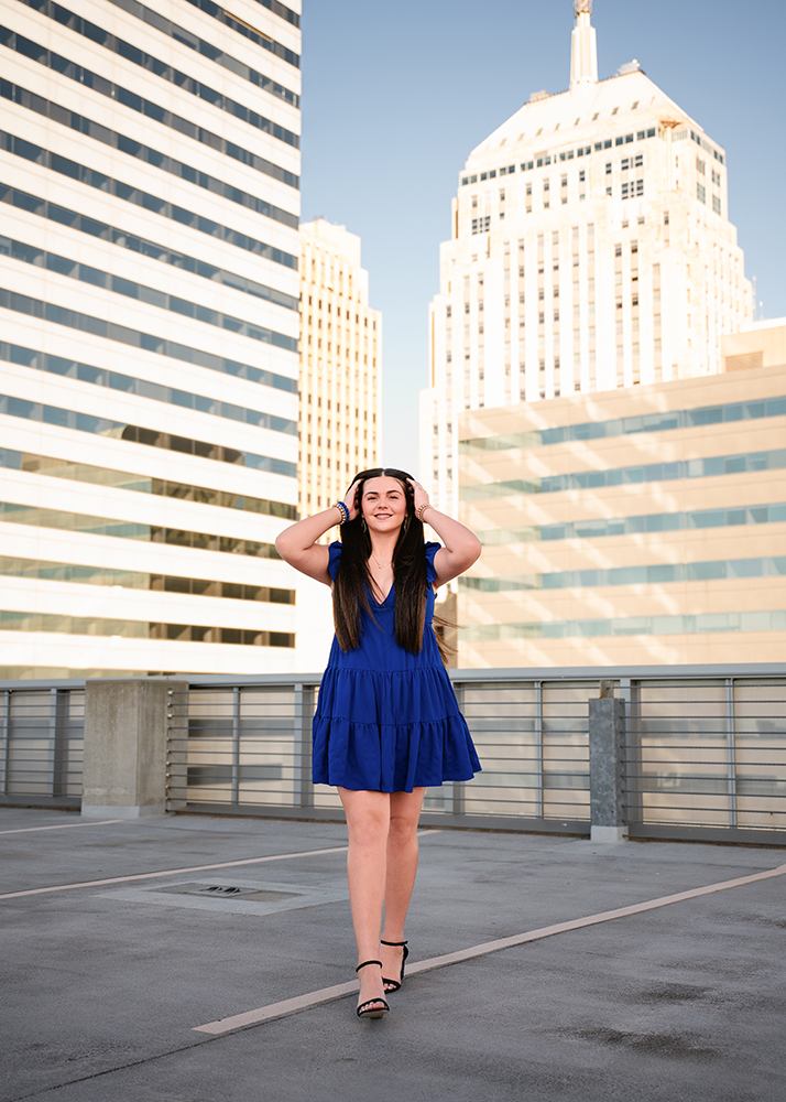 A senior girl in a blue dress stands confidently on a rooftop with a city skyline of tall buildings in the background, under a clear blue sky.