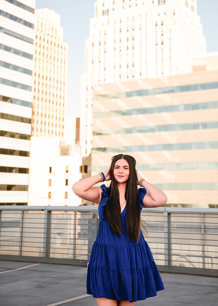 Senior girl in a blue dress walks toward the camera on a rooftop with modern city buildings in the background. She smiles, exuding confidence and joy.