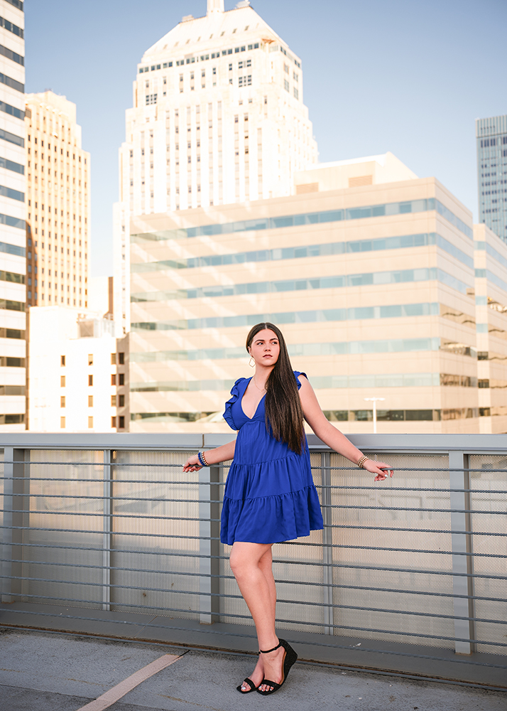 A senior girl in a royal blue dress stands confidently on a rooftop, framed by city skyscrapers under a clear sky. She looks serene and composed.
