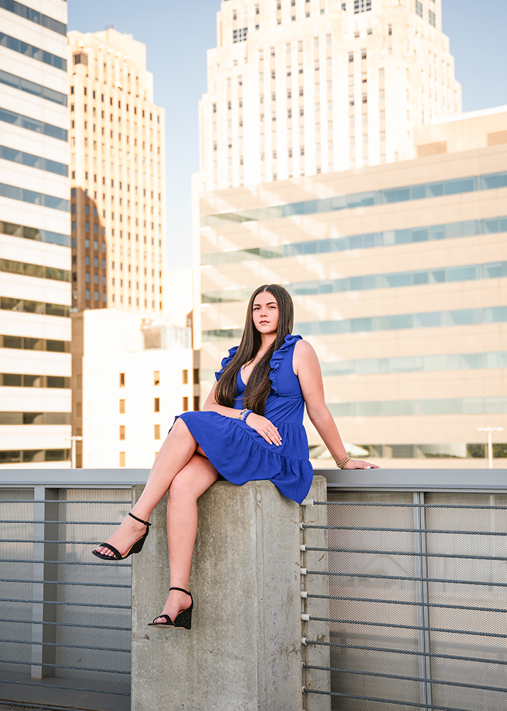 A senior girl in a blue dress sits on a concrete ledge of a modern building, with skyscrapers in the background. The scene is bright and urban.