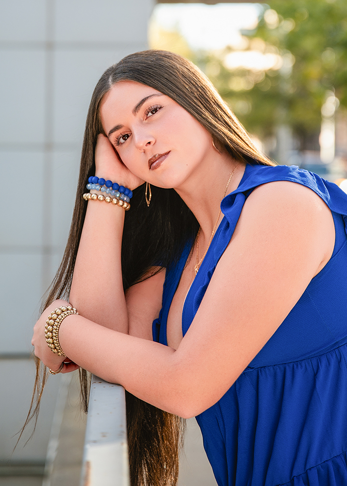 High school senior in a blue dress leans on a railing with a calm expression. She wears bracelets and long earrings, framed by soft, sunny lighting and greenery.
