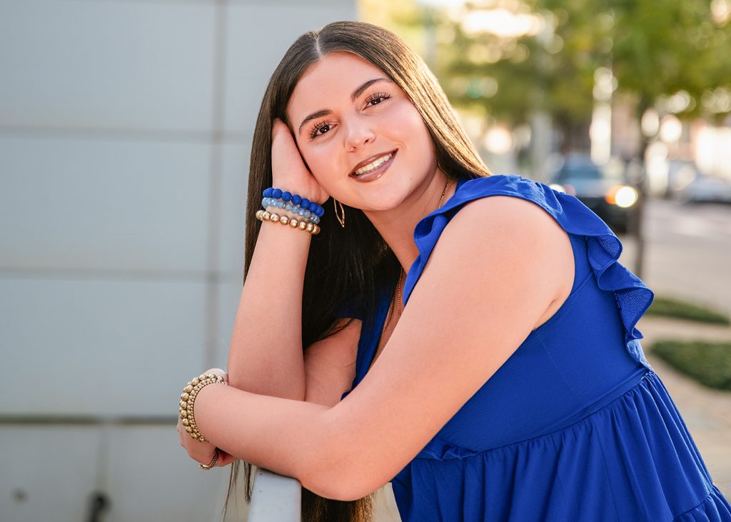 A senior girl in a bright blue dress leans on a railing, smiling warmly. They have long brown hair and wear gold and blue bracelets. The background shows a blurred urban setting.