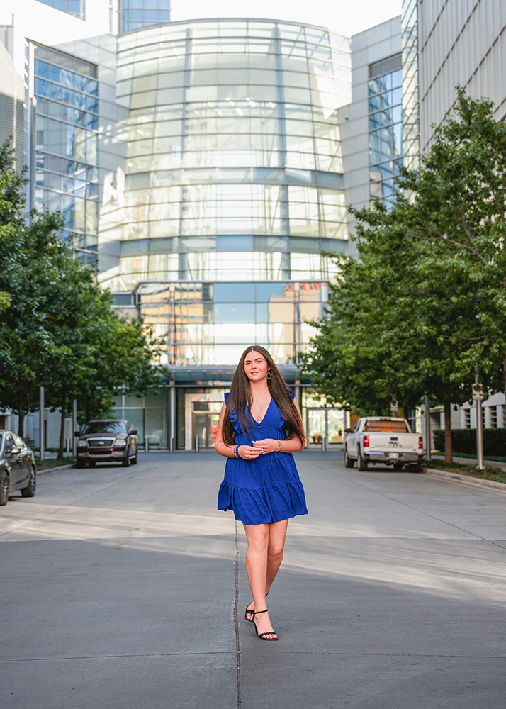 Senior girl in a blue dress walks confidently down an urban street flanked by greenery and parked cars, against a backdrop of modern glass architecture.