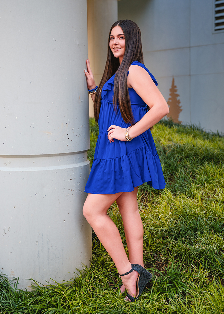 Peyton poses for her downtown OKC senior photo session in a vibrant blue dress leans against a white column, smiling. She stands on lush green grass, wearing sparkly heels. The mood is cheerful and relaxed.