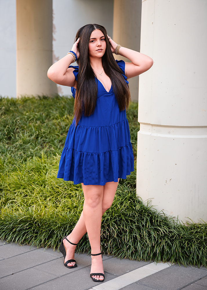 Senior girl in a flowing blue dress and black heels stands confidently with hands in long, dark hair against a background of grass and concrete pillars.