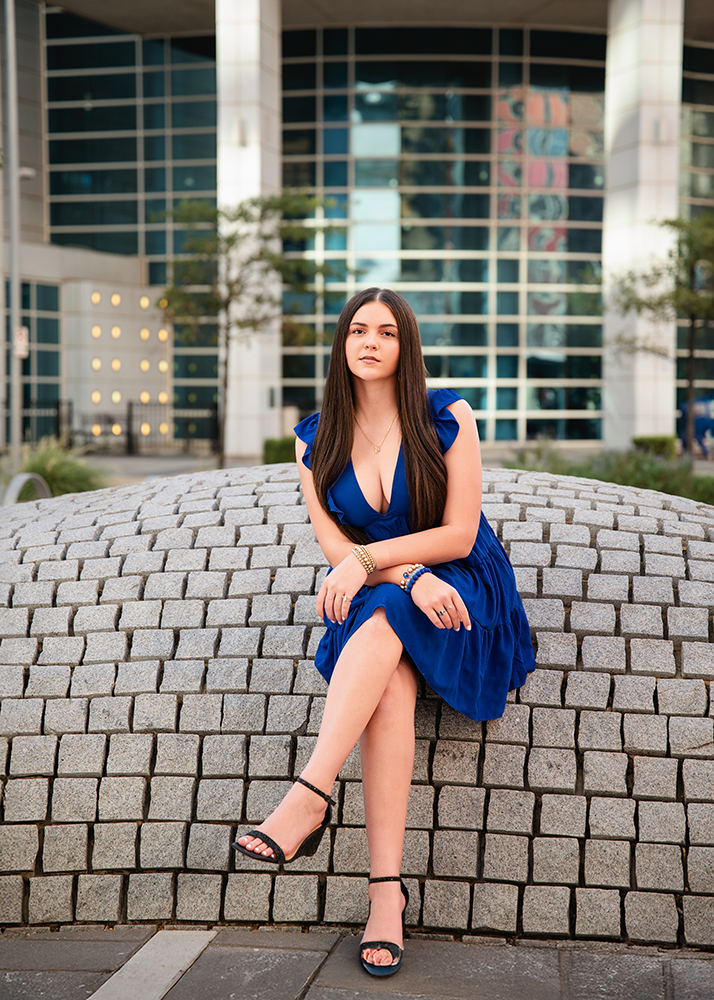 Senior girl in a blue dress sits confidently on a curved gray stone structure. Behind her is the downtown OKC library with large windows and trees, creating an urban, stylish atmosphere.