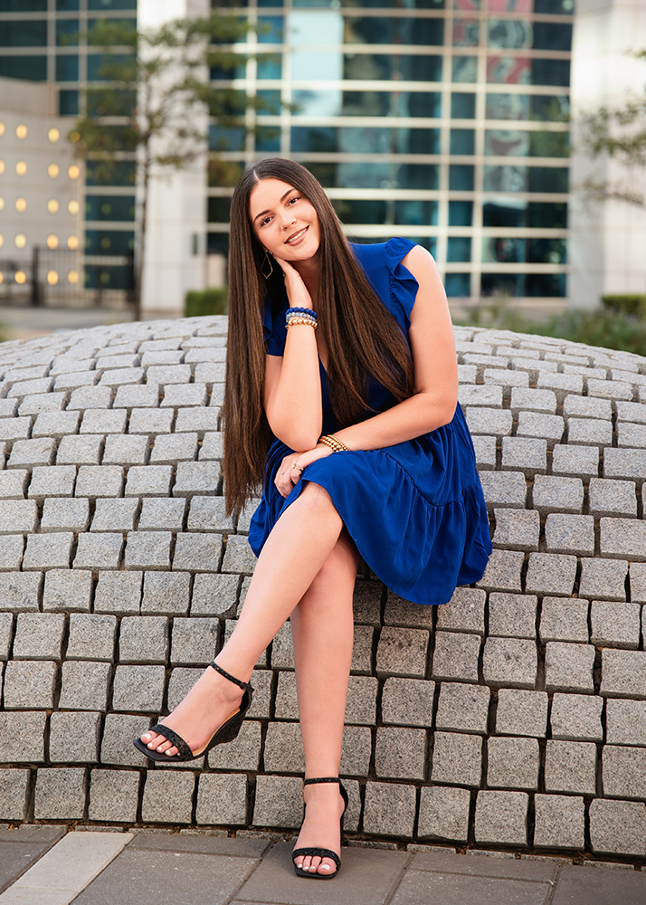 A high school senior in a blue dress sits smiling on a cobblestone wall, set against a modern building. Her relaxed pose conveys a cheerful, confident tone.