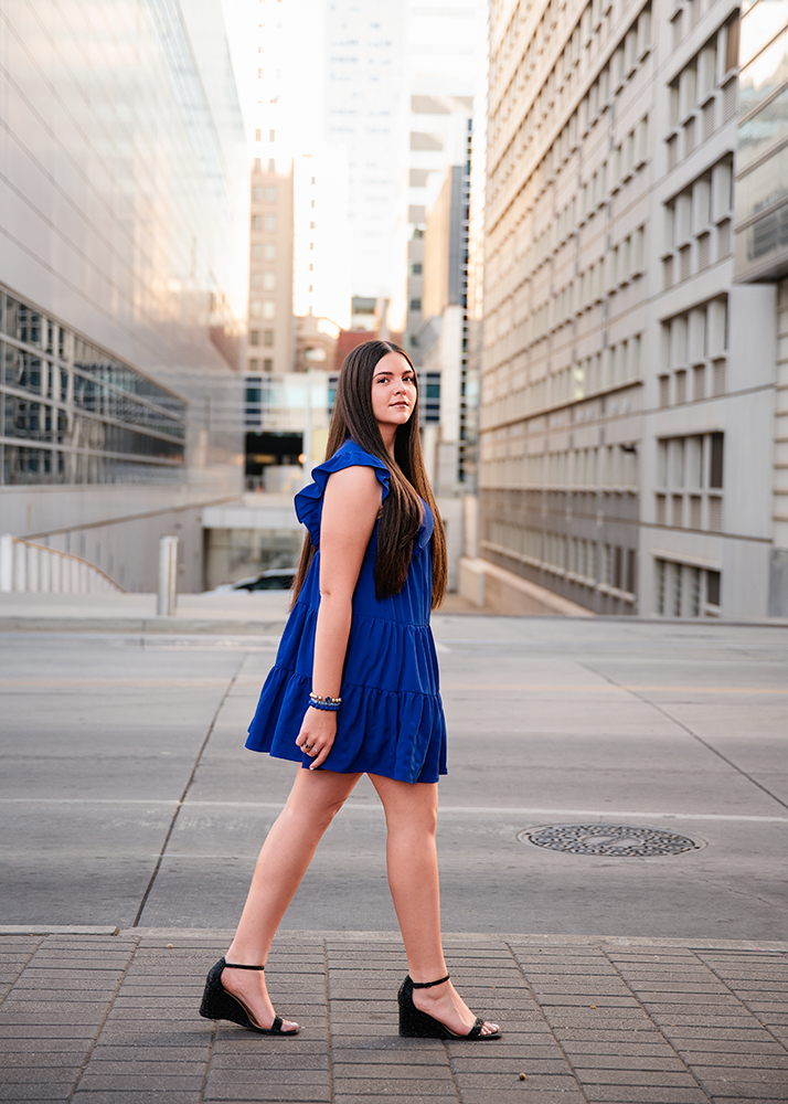 A senior girl in a blue dress and black wedges walks confidently through a city street lined with tall buildings, exuding a calm and focused demeanor.