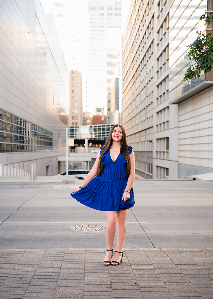 A senior girl in a blue dress and black wedges walks confidently through a city street lined with tall buildings, exuding a calm and focused demeanor.