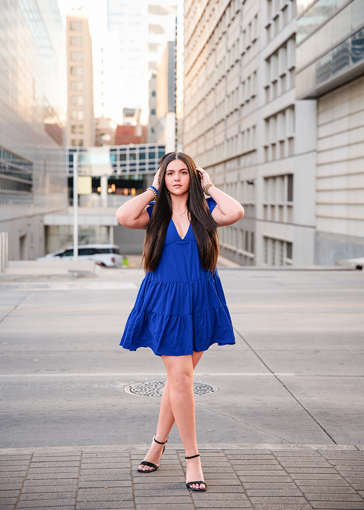 A senior girl in a vibrant blue dress smiles while standing on a city street. She holds the dress slightly out to the side. The background features modern buildings and a clear sky, creating a bright and cheerful urban scene.