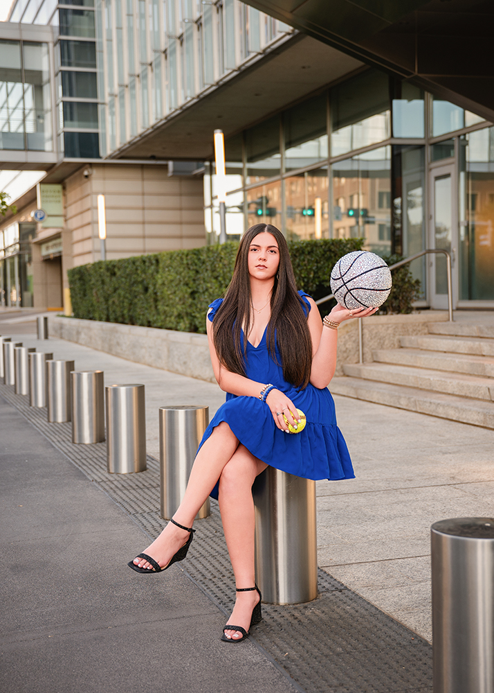 A senior girl in a blue dress sits on a metal bollard, holding a sparkling basketball. She appears confident, set against a modern urban building backdrop.