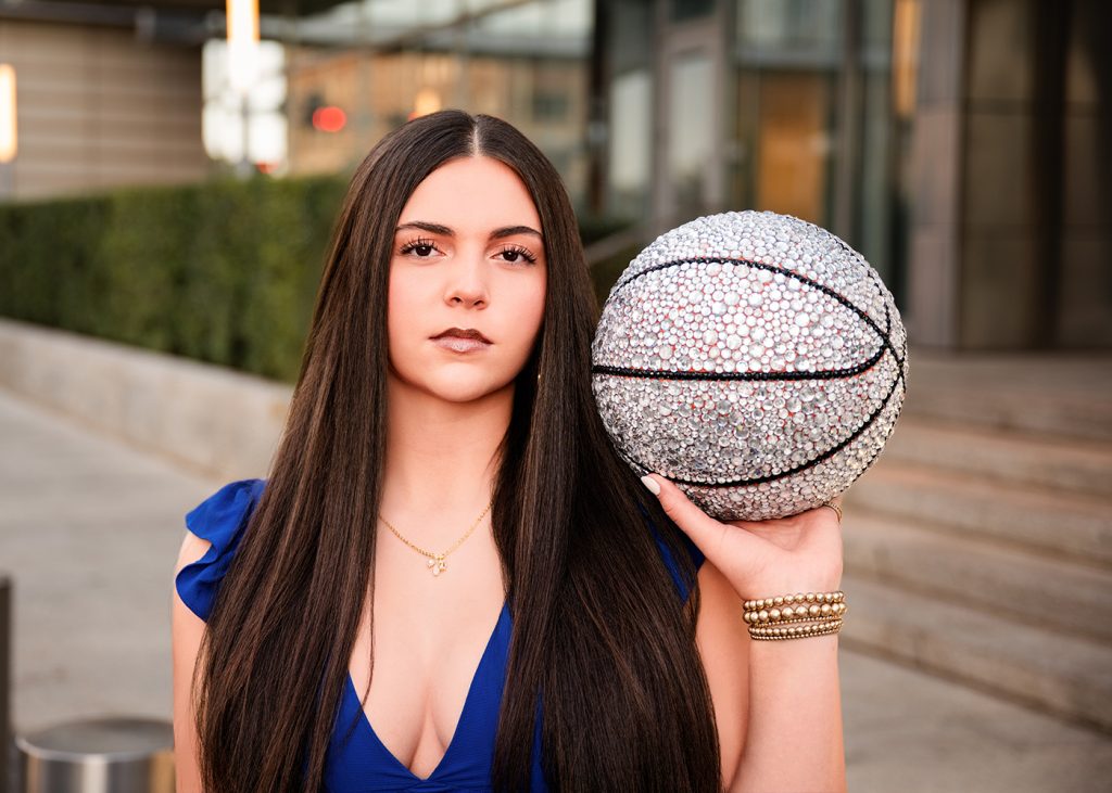 A high school senior with long dark hair holds a rhinestone-studded basketball, wearing a blue dress and gold bracelet. She stands confidently outside, evoking elegance.