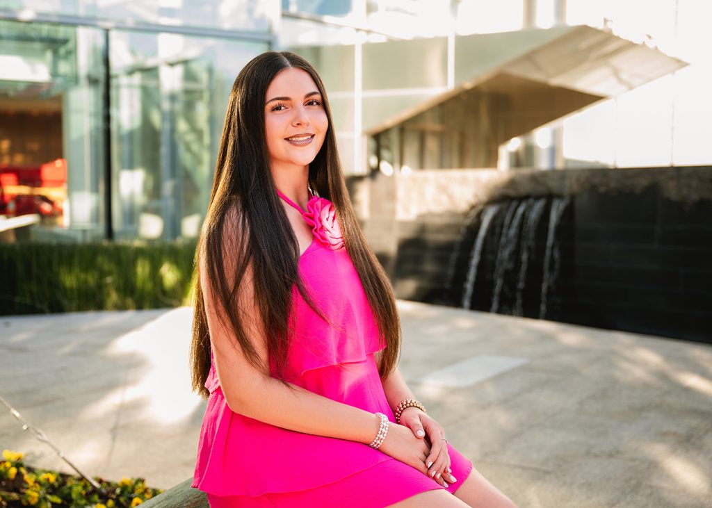 A woman with long dark hair, smiling and seated in front of the Devon Tower in downtown Oklahoma City. She wears a bright pink dress and bracelets. A water feature is in the background, under soft sunlight.