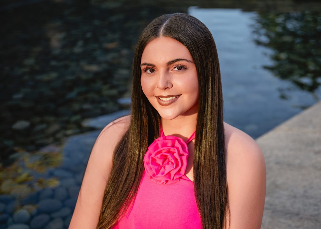 A high school senior with long brown hair smiles warmly, wearing a pink top with a large flower. She is sitting by a calm pool, creating a serene scene.