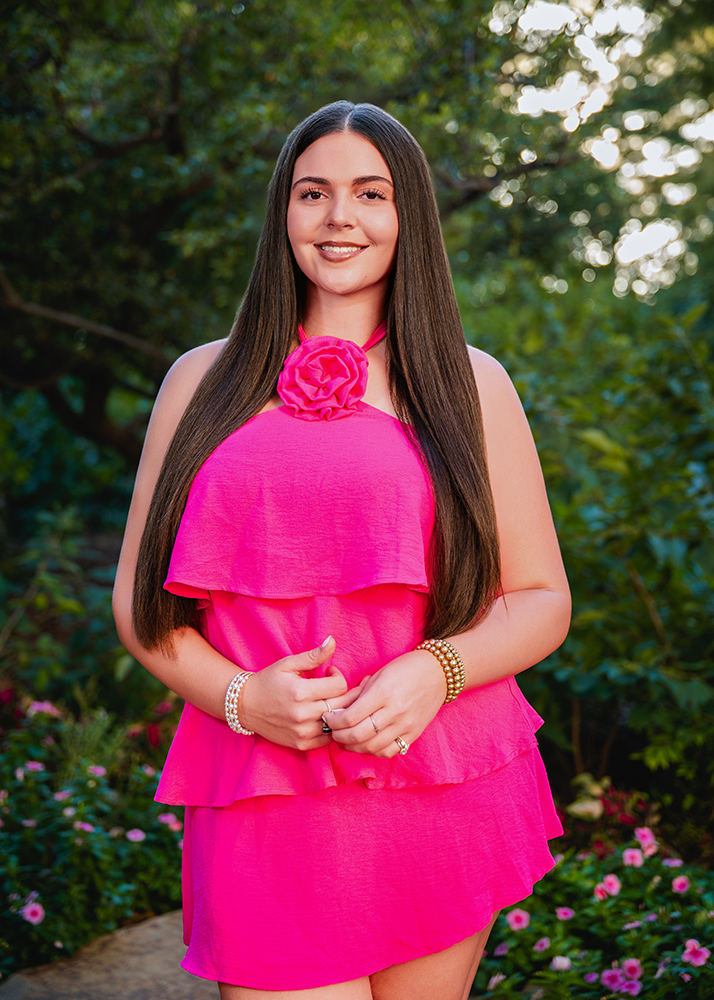 A high school senior in a bright pink dress with a flower detail stands smiling in a green garden. Her long hair is straight, and she wears pearl bracelets, conveying a cheerful and vibrant mood.