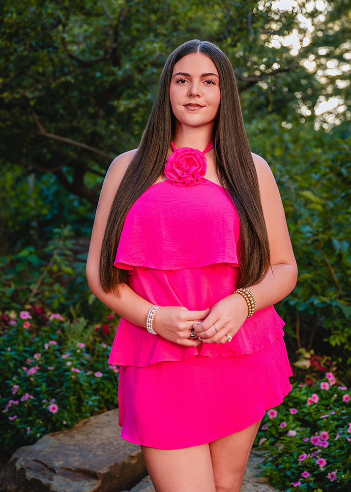 A high school senior in a bright pink dress with a flower detail stands smiling in a green garden. Her long hair is straight, and she wears pearl bracelets, conveying a cheerful and vibrant mood.