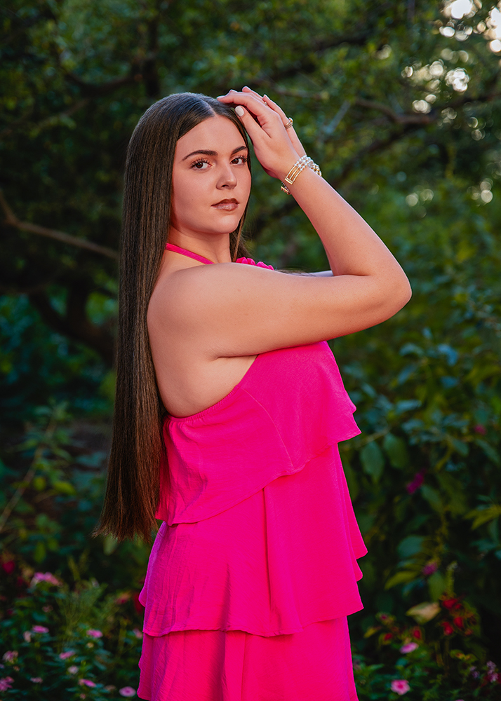 Peyton posing for her senior photos in a bright pink dress with a floral detail, outdoors in a garden setting. She has long straight hair and is wearing bracelets, exuding confidence.