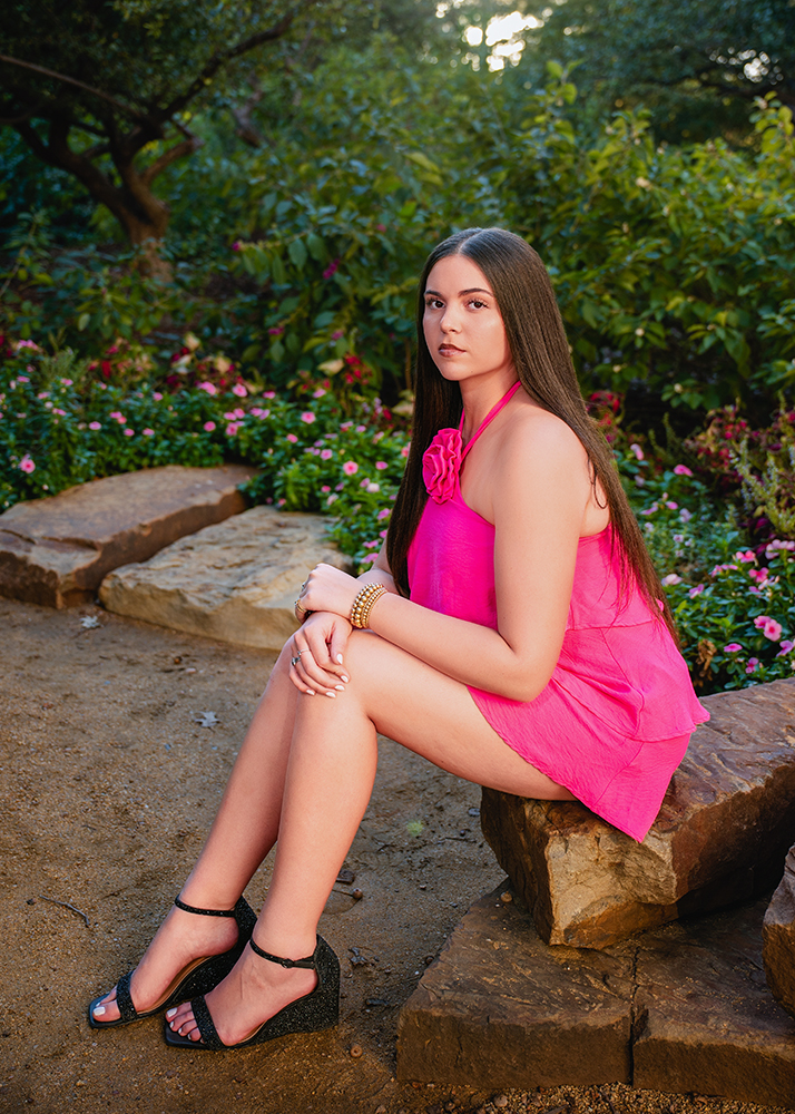 A senior girl in a bright pink dress sits on a stone bench in a garden, surrounded by lush greenery and flowers. She looks calmly at the camera.