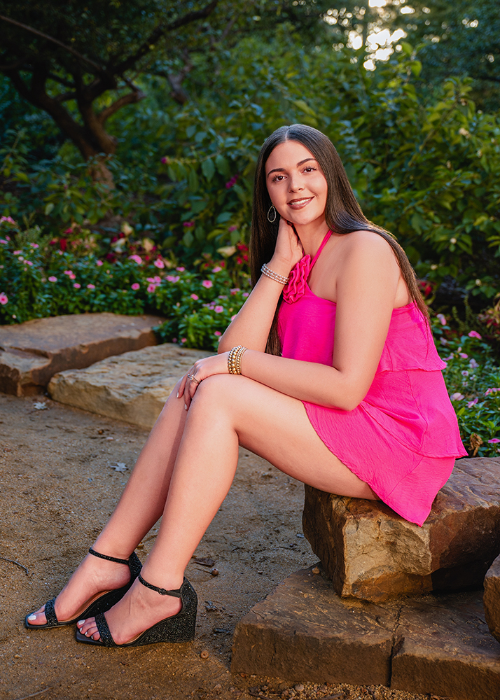 A senior girl in a pink dress sits on a stone, surrounded by lush green foliage and vibrant flowers. She exudes a calm demeanor, looking thoughtfully at the camera.