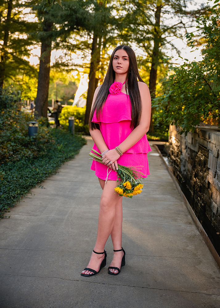 A senior girl in a pink dress sits on a stone, surrounded by lush green foliage and vibrant flowers. She exudes a calm demeanor, looking thoughtfully at the camera. in a bright pink dress holds sunflowers, standing on a garden path. Trees and greenery surround her, with sunlight filtering through.