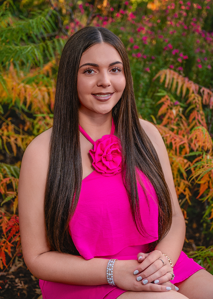 High school senior in a vibrant pink dress with a rose detail smiles warmly. She has long, straight hair and sits among lush greenery and pink flowers.