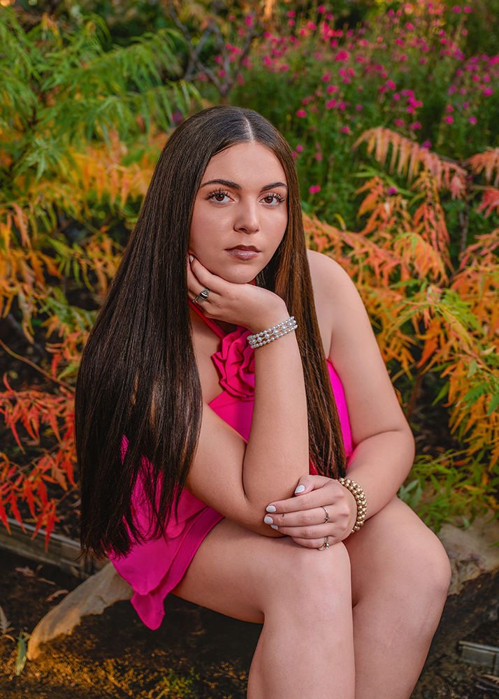 High school senior in a vibrant pink dress with a rose detail smiles warmly. She has long, straight hair and sits among lush greenery and pink flowers.