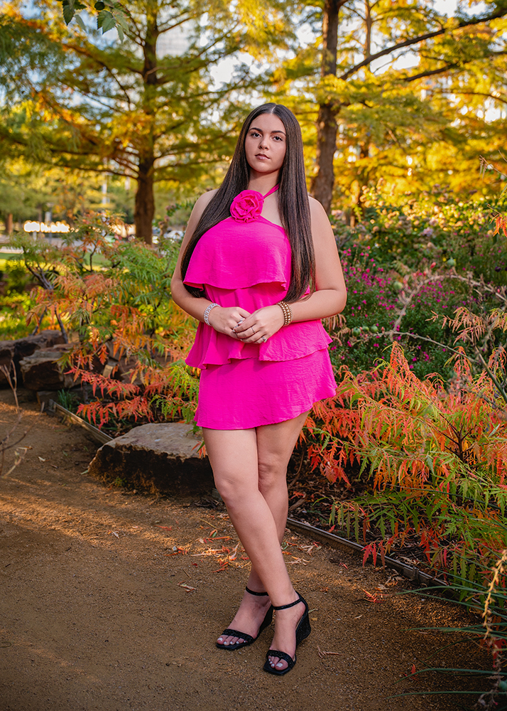 A high school senior in a vibrant pink dress poses confidently in a garden. Green and orange foliage surrounds her, with soft evening light creating a serene, warm atmosphere.
