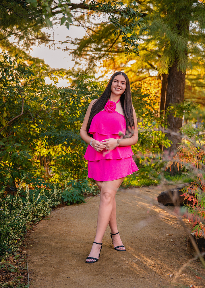 A high school senior in a vibrant pink dress poses confidently in a garden. Green and orange foliage surrounds her, with soft evening light creating a serene, warm atmosphere.