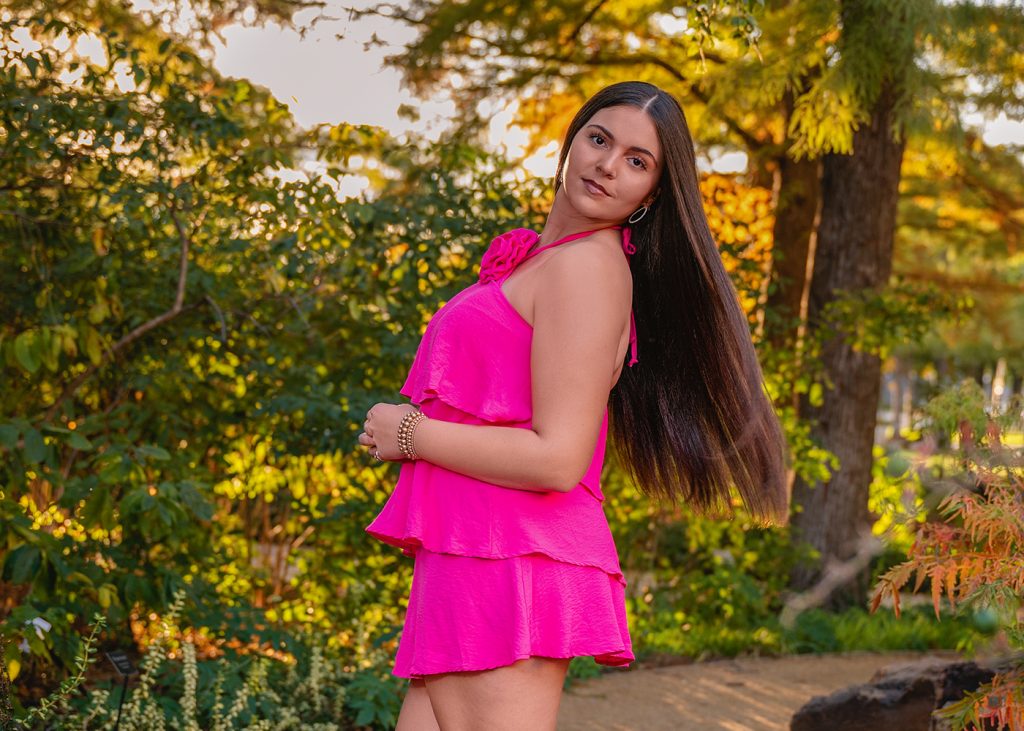 A high school senior in a vibrant pink dress stands on a garden path, surrounded by lush green foliage. Her expression is serene beneath a sunset glow.
