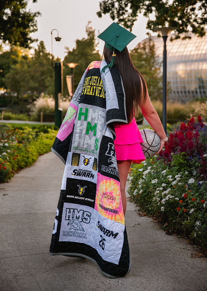 A graduate in a bright pink dress walks away on a garden path, wearing a green cap and gown draped over her shoulder. She holds a basketball.