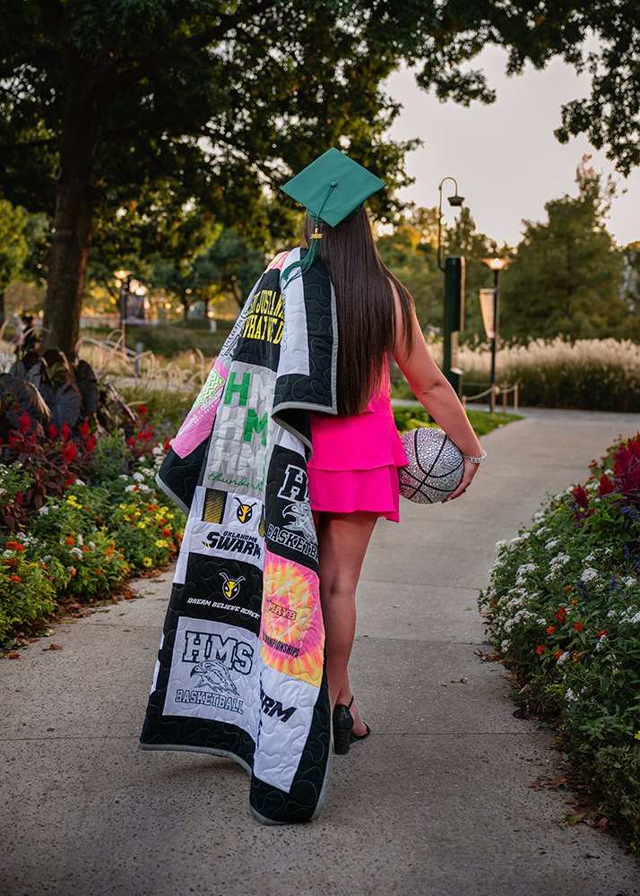 A young woman in a pink dress and green graduation cap walks along a garden path. She drapes a quilt with various patterns over her shoulder and holds a glittery basketball, symbolizing achievement and nostalgia.