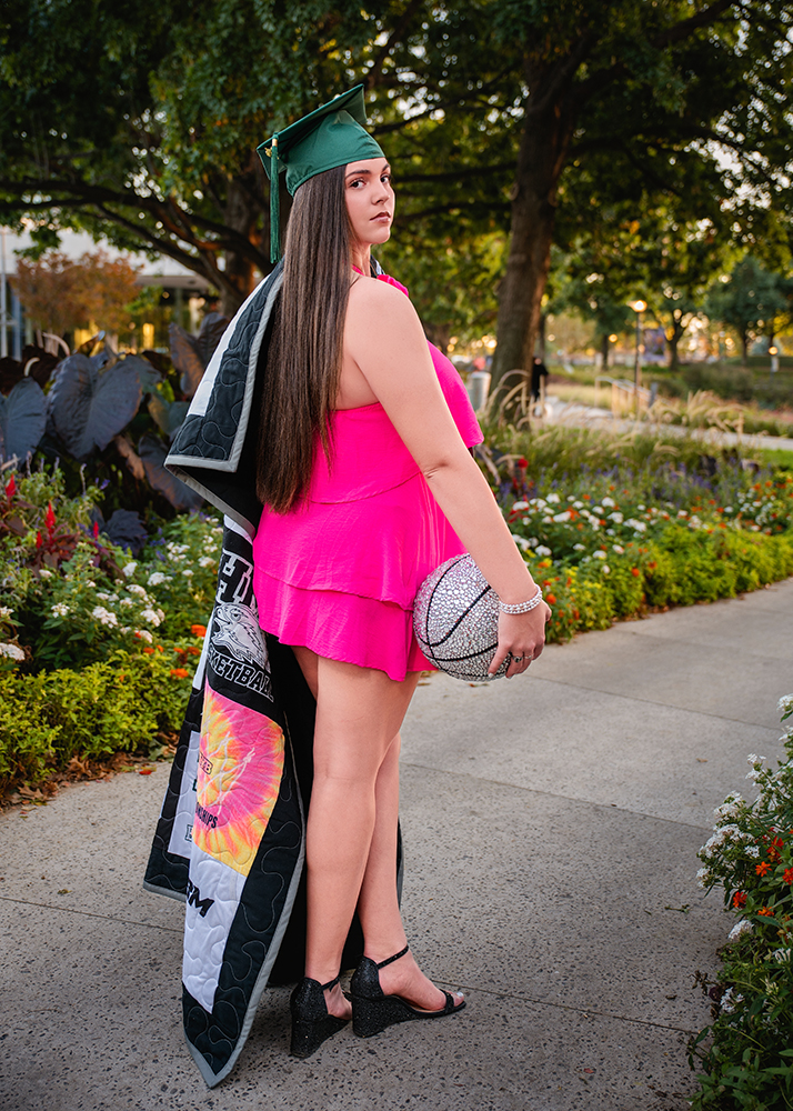 A high school senior in a bright pink dress and green graduation cap stands on a garden path, holding a silver basketball and a quilt draped over her shoulder.