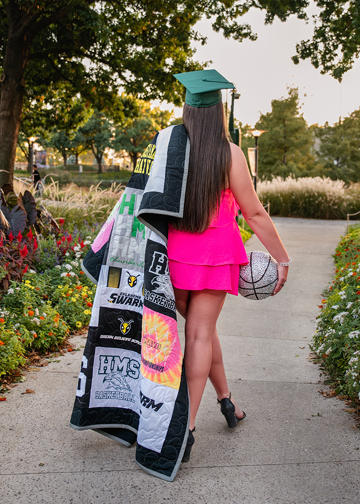 Young woman in a green graduation cap, pink dress, holding a quilt and sparkly basketball, walks on a scenic path lined with flowers and trees.