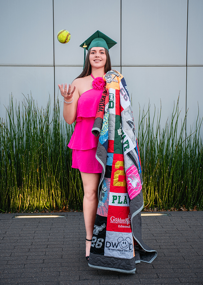 Young woman in a green graduation cap and pink dress, smiling, juggles a softball. She holds a colorful quilt while standing in front of tall grass.