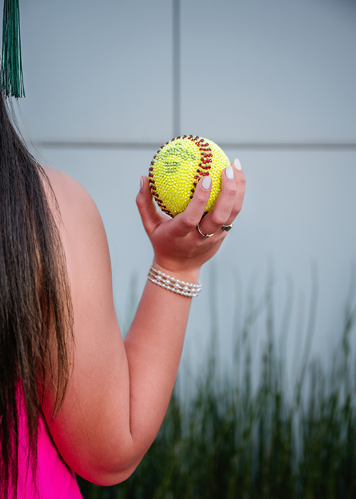 A person in a pink top holds a sparkling, gem-studded yellow softball. A green tassel hangs nearby, suggesting graduation. The background is blurred greenery.