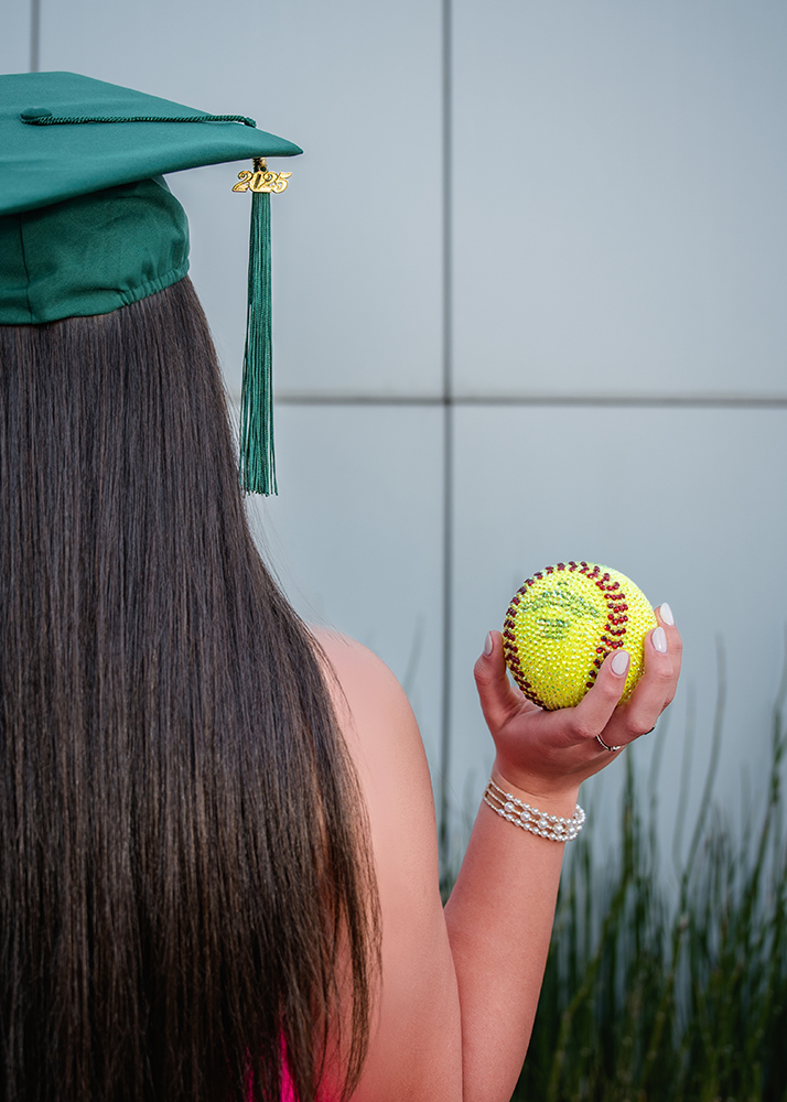 A person in a pink top holds a sparkling, gem-studded yellow softball. A green tassel hangs nearby, suggesting graduation. The background is blurred greenery.