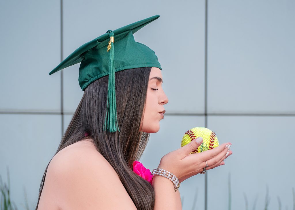 Edmond Santa Fe senior in a green graduation cap, eyes closed, holds a bedazzled softball, creating a moment of reflection and achievement against a grey background.