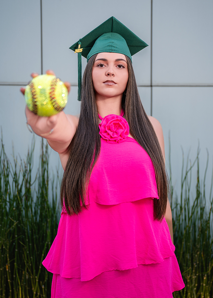 A young woman in a vibrant pink dress and green graduation cap extends a softball toward the camera, standing confidently against a modern backdrop.