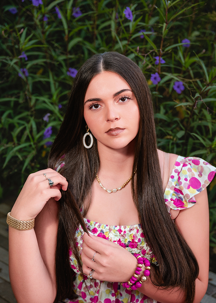 A young woman with long dark hair stands outdoors, wearing floral attire, gold jewelry, and pink bracelets against a backdrop of green foliage and purple flowers.