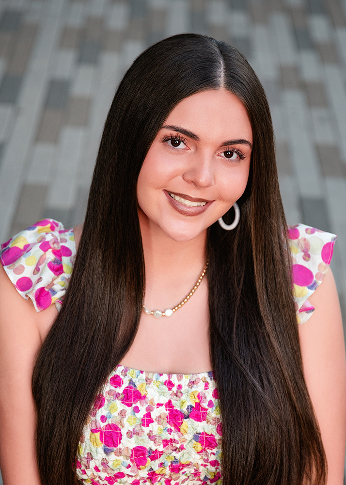 Smiling high school senior with long, straight hair, wearing a colorful floral top and hoop earrings. Background features blurred stone tiles, conveying a cheerful tone.