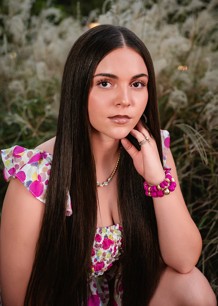 High school senior with long dark hair kneels in a field of pampas grass, wearing a floral dress and vibrant pink bracelets. She gazes at the camera with a serene expression.