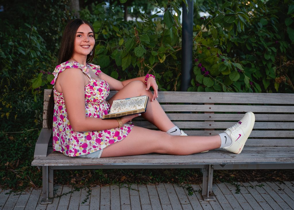 A high school senior in a floral dress smiles while sitting on a park bench, holding an open book. Her relaxed pose and lush greenery convey a peaceful mood.