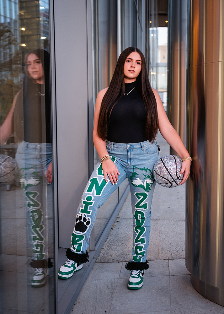 A young woman stands confidently by a glass wall, holding a glittery basketball. She wears a black top and jeans with "SENIOR 2025" and a wolf design. Her reflection is visible in the glass, adding depth. The mood is empowering and stylish.