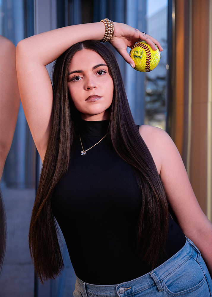 A high school senior with long dark hair poses confidently, holding a decorated softball. They're wearing a black top and jeans, exuding a calm, focused demeanor.