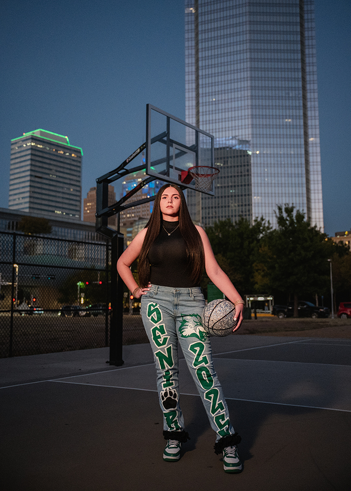 Senior girl standing confidently on an outdoor basketball court at dusk, holding a basketball. Her jeans say "Senior 2025" in green lettering.