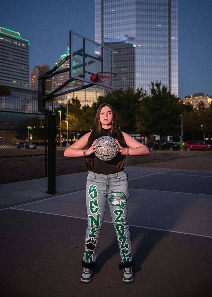 Senior girl stands on an outdoor basketball court at dusk, holding a glittery ball. She wears jeans with "Senior 2024" text. Urban skyline in background.