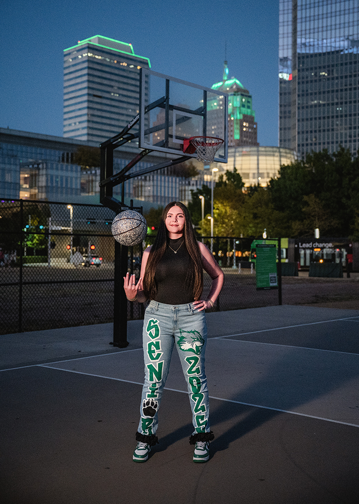 Senior girl stands on a basketball court, balancing a ball on her fingertip. She wears jeans with "Senior 2023." Cityscape with lit buildings in the background.