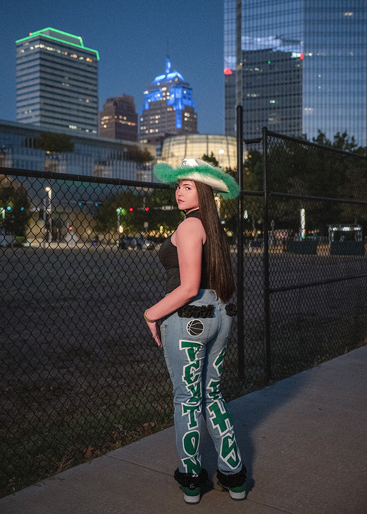 Senior girl in a green cowboy hat and jeans with bold lettering stands on a sidewalk at dusk. Oklahoma City skyscrapers with neon lights shimmer in the background.